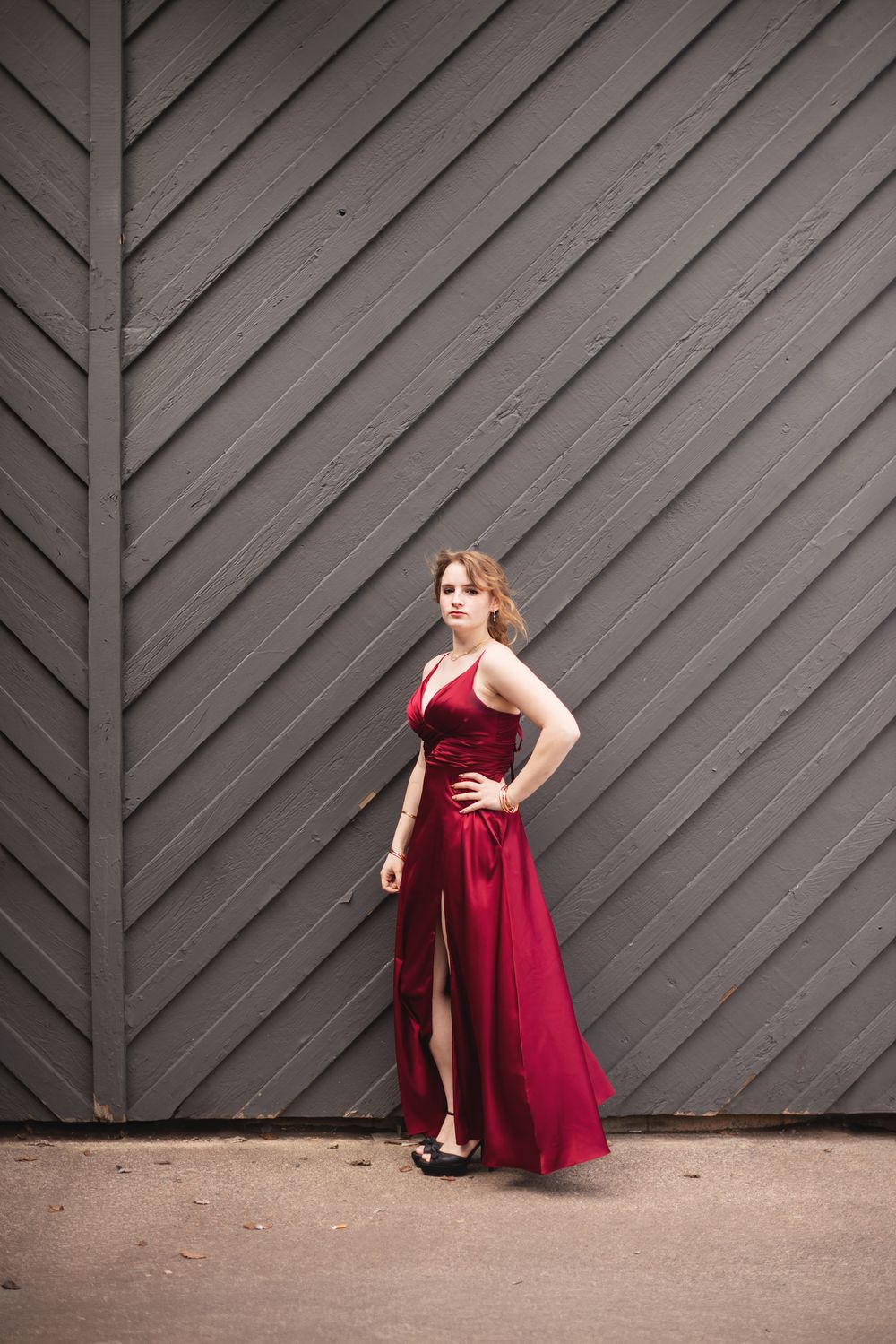 A young woman in a flowing red satin gown poses confidently against a gray chevron wooden wall backdrop.