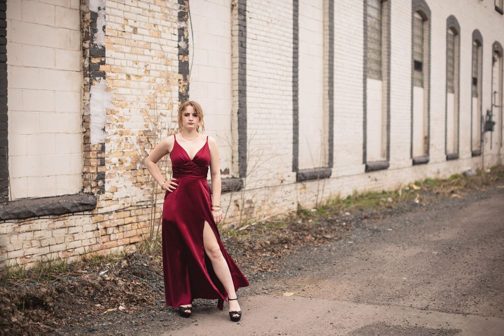 Woman in burgundy slit gown posing beside weathered white brick building on urban street.