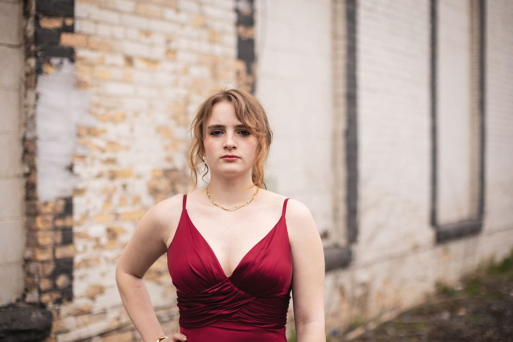 Young woman in elegant red spaghetti strap dress posing confidently against rustic brick wall backdrop.