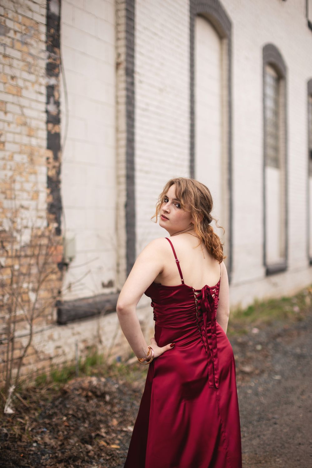 Young woman in elegant red satin gown with lace-up back poses outdoors near rustic brick building.