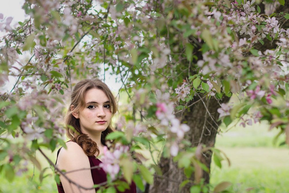 Young woman posing among pink blossoming tree branches outdoors in a lush green garden setting.