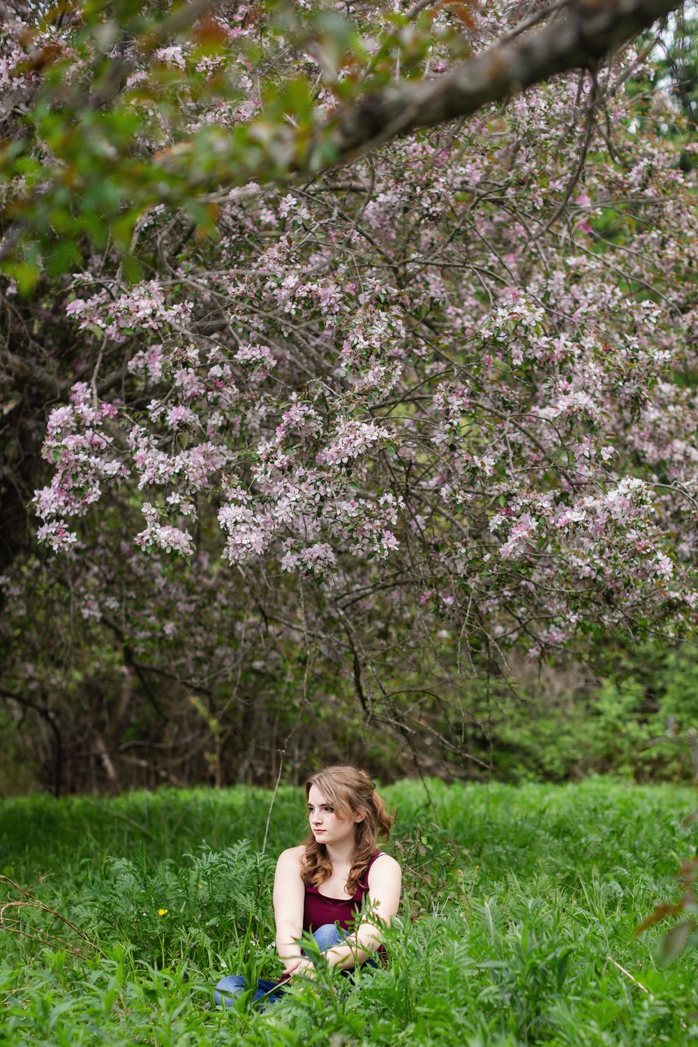 Young woman sitting in lush green grass beneath a blooming pink cherry blossom tree in spring.