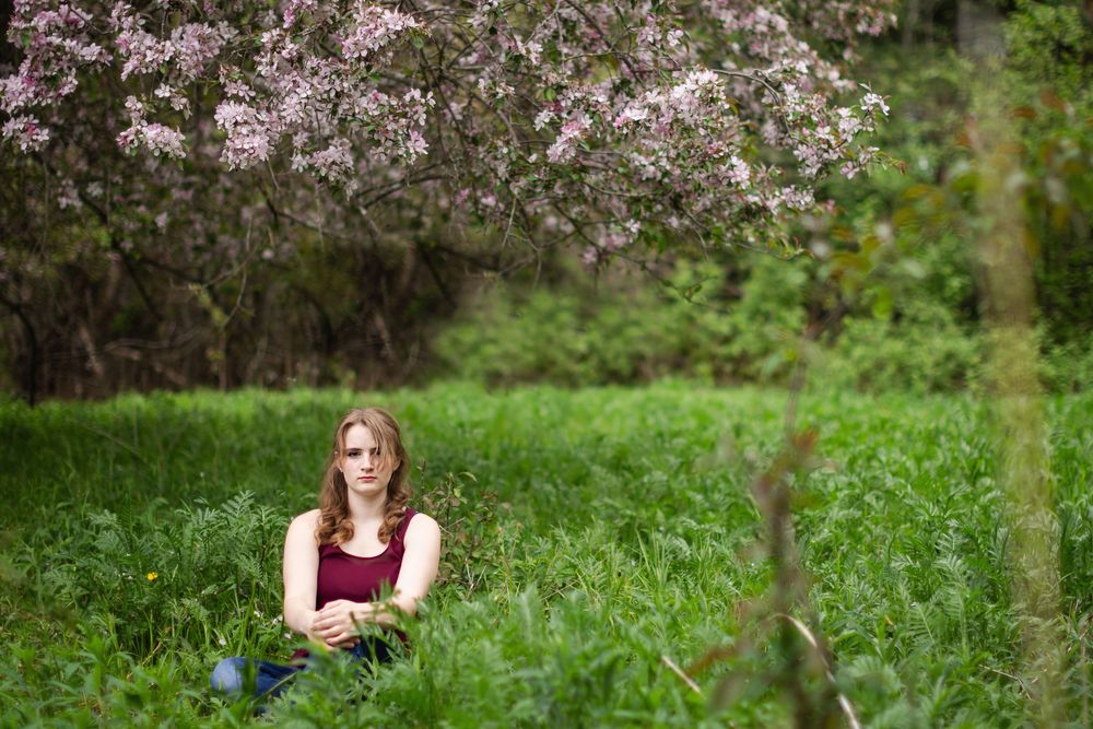 Young woman sitting in lush green grass beneath blooming pink flowering tree in serene outdoor setting.