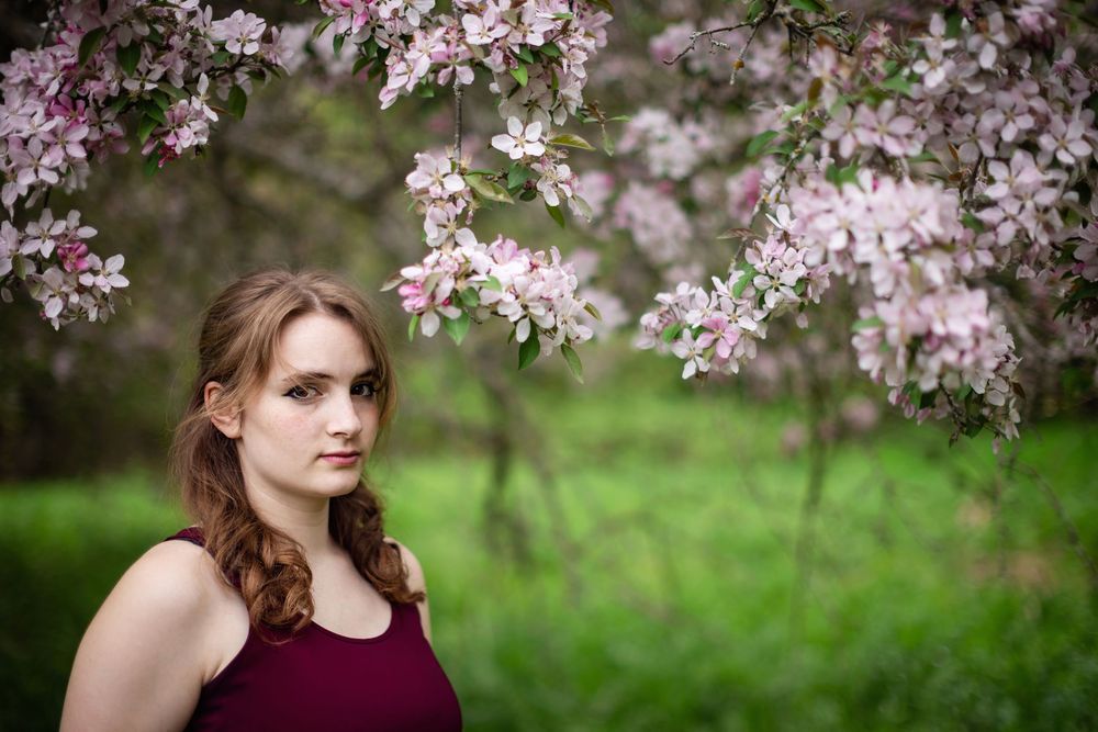Young woman in burgundy top poses under blooming pink cherry blossom tree branches in a lush green garden.