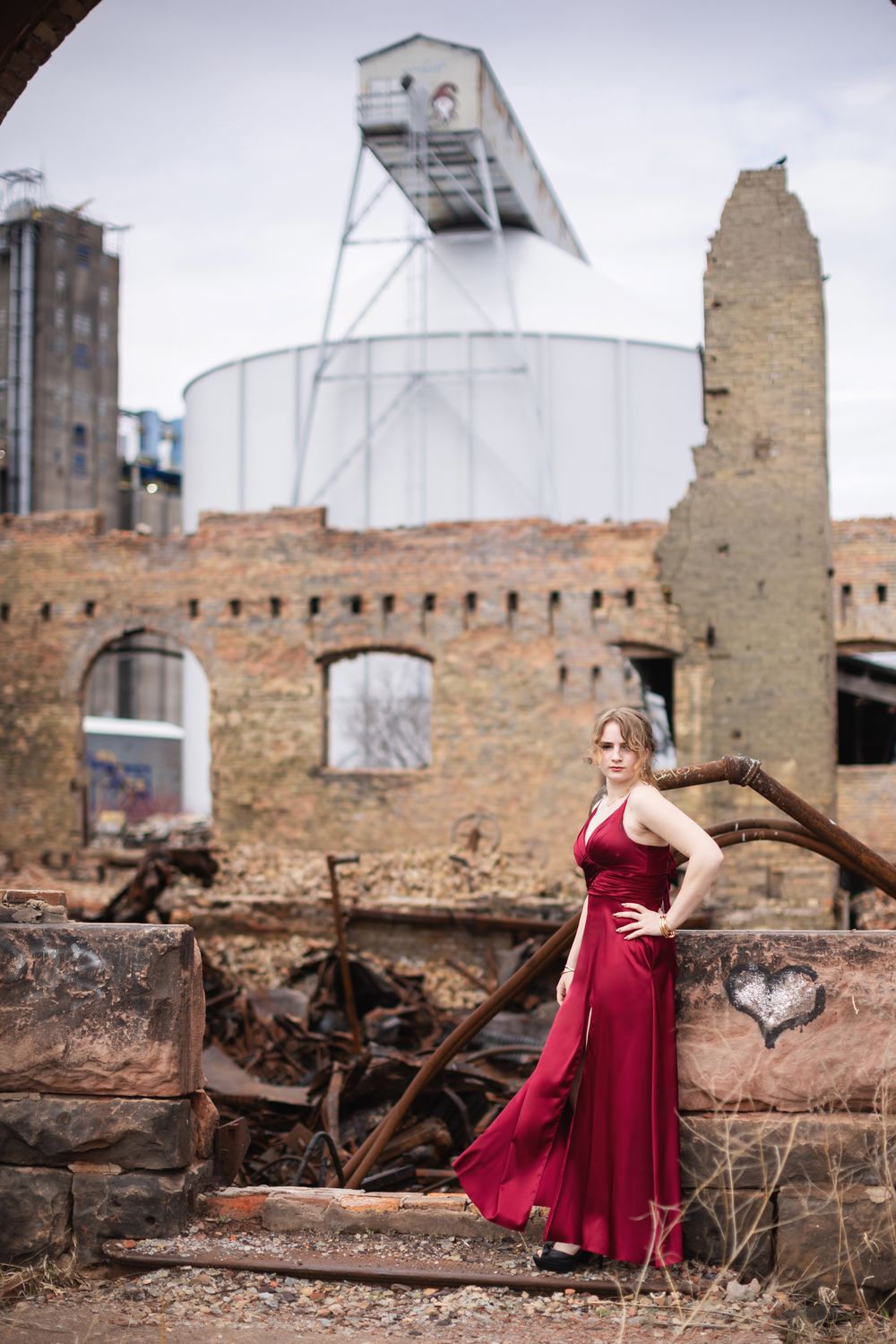 Elegant woman in red gown posing before industrial ruins with brick walls and metal structures.