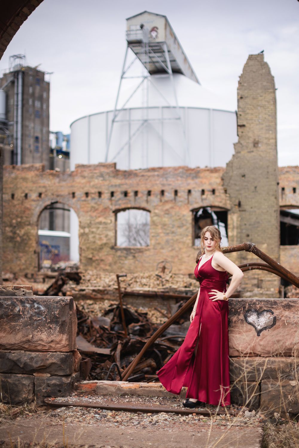 Woman in red dress poses gracefully among rustic ruins and industrial structures under an overcast sky.