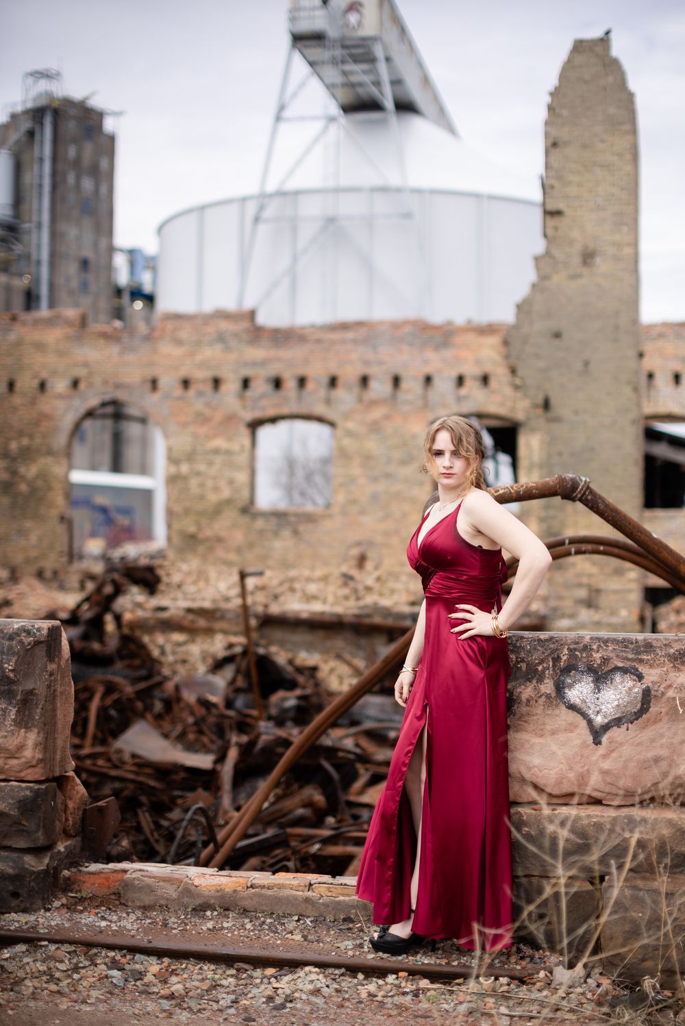 Woman in elegant red gown posing at rustic industrial ruins with brick walls and metal structures.