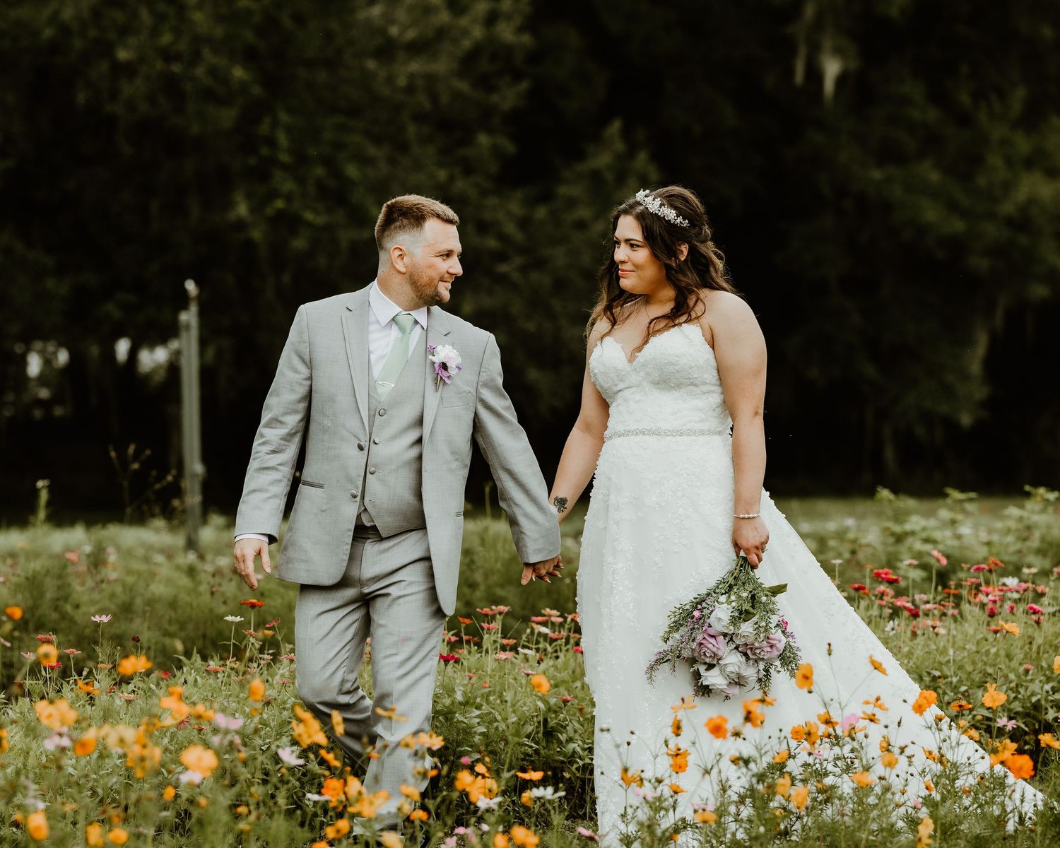 Bride and groom walking hand in hand through a vibrant wildflower meadow, smiling at each other on their wedding day.