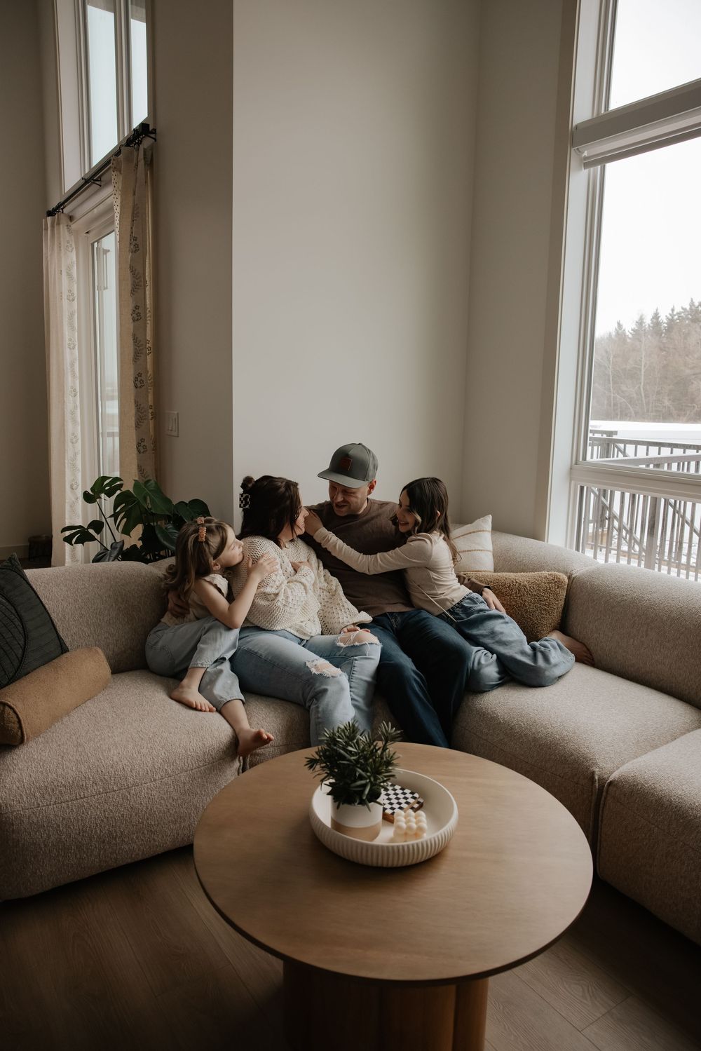 A family of four sits together on a cozy sectional sofa, sharing a warm embrace in a bright, modern living room.