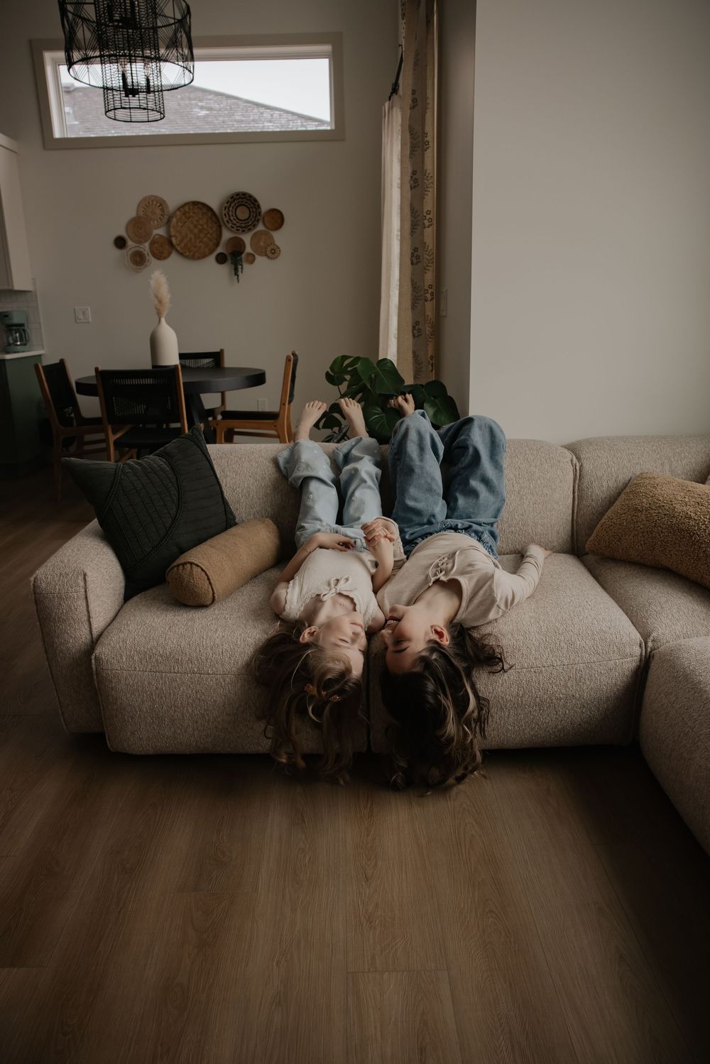 Two children lying upside down on a beige couch in a modern living room with wood floors.