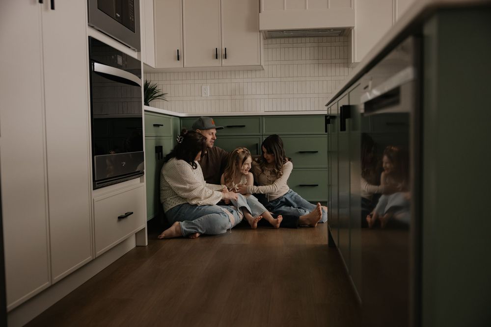 Family sitting together on kitchen floor laughing, surrounded by green cabinets and modern appliances.