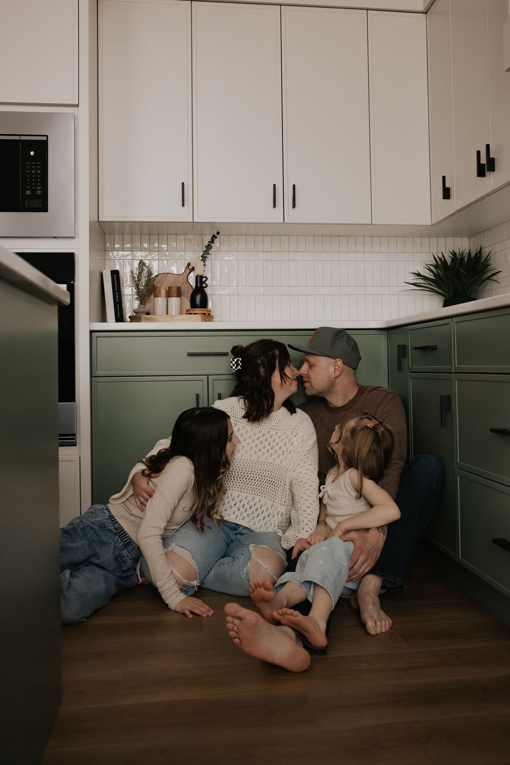 Family of four sitting on kitchen floor, parents kissing, surrounded by children in green cabinet kitchen.