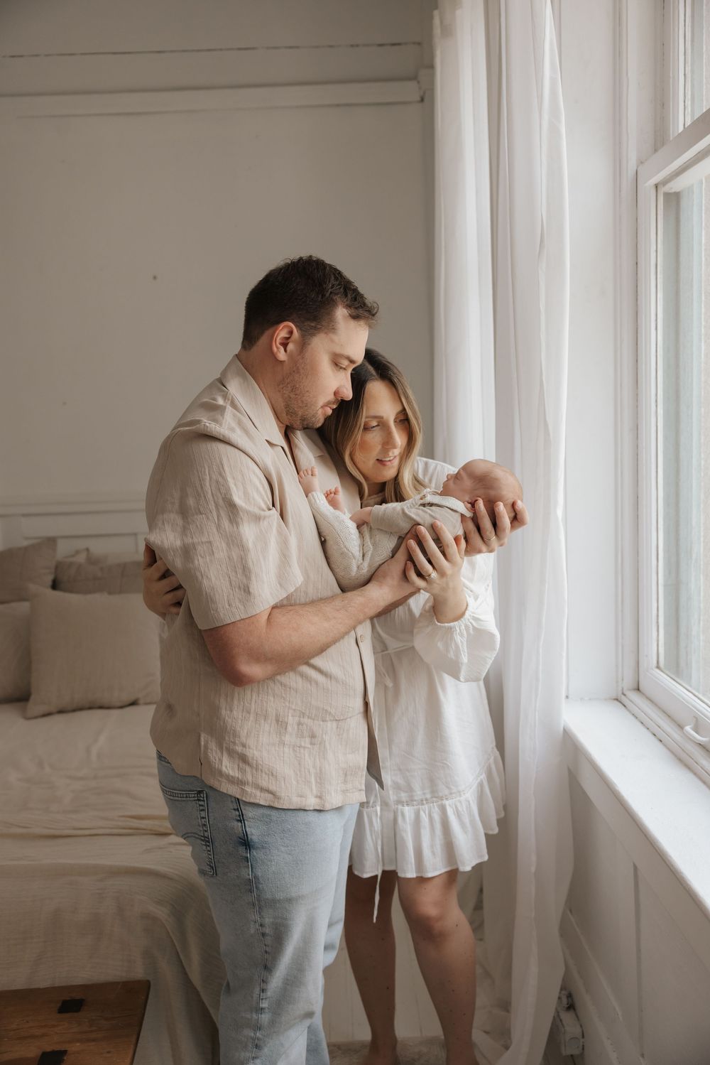 A couple stands by a window in a bright bedroom, lovingly cradling their newborn baby together.