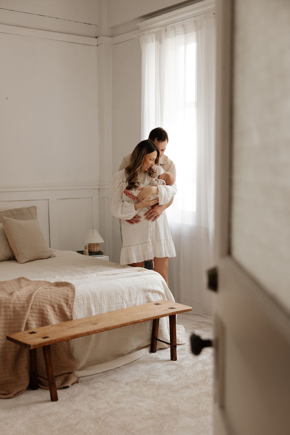 A couple in a bright bedroom holds their newborn baby near a window, dressed in white, with a wooden bench and bed nearby.