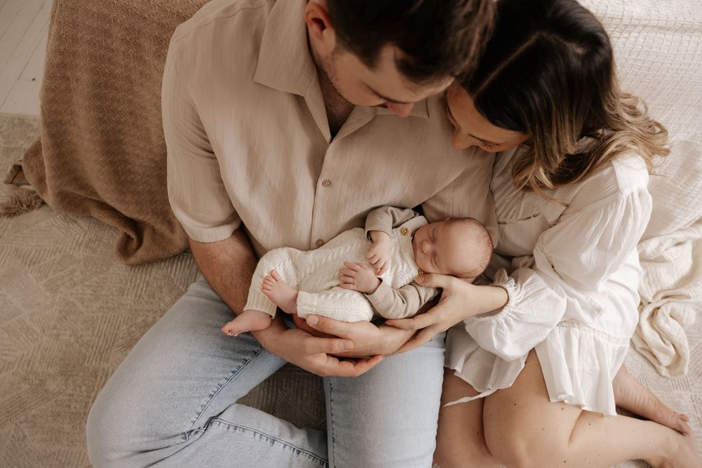 Parents sitting on floor gazing lovingly at their newborn baby wrapped in white knit outfit during professional photoshoot.