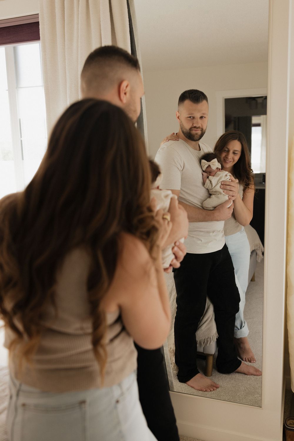 A couple holding their newborn baby admires their reflection in a large mirror in a bright bedroom.