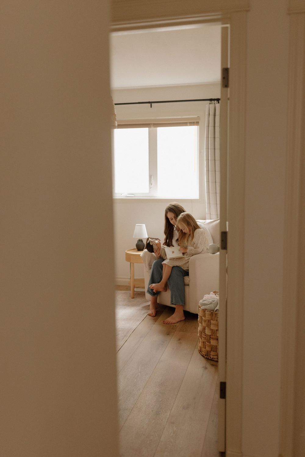 Two women sitting together on a couch in a warmly lit bedroom with hardwood floors, viewed through a doorway.