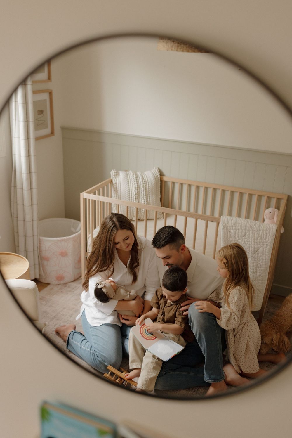 Family of four with newborn baby reflected in round mirror, sitting on nursery floor beside wooden crib.
