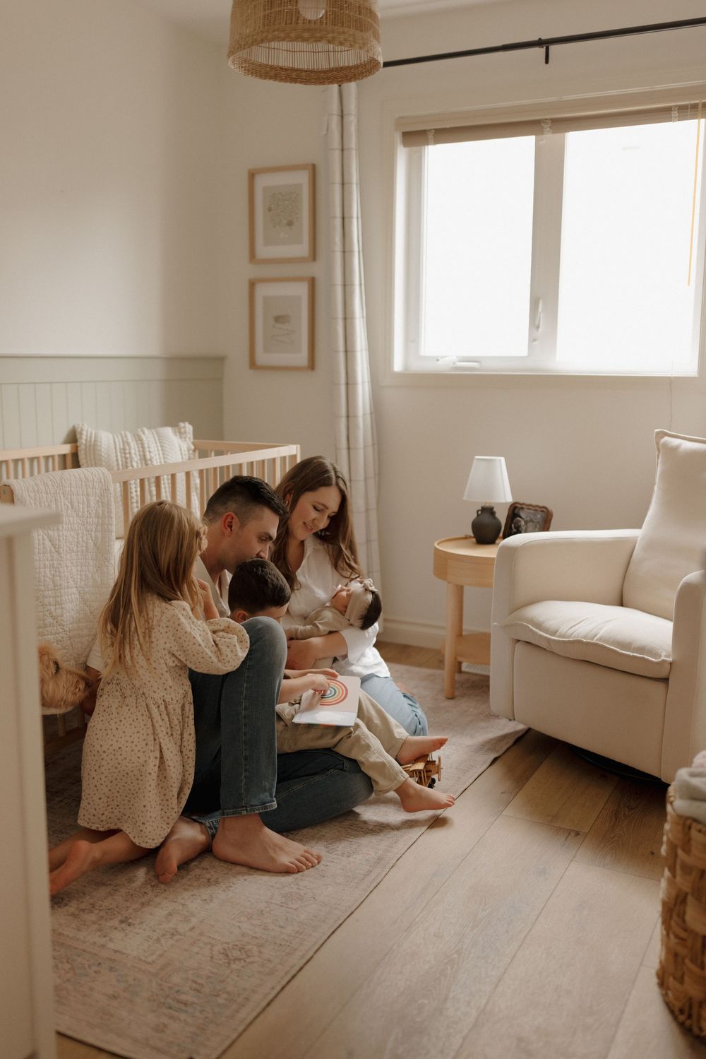 A family of five sits together on a nursery floor, with parents and children gathered around a newborn baby.