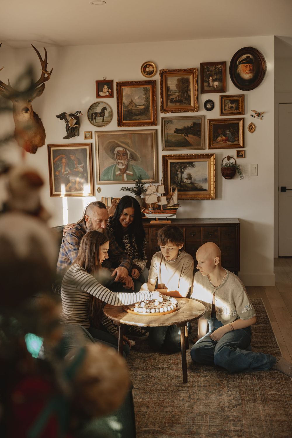 Family gathered around a small table playing a board game in a cozy living room with vintage art gallery wall.