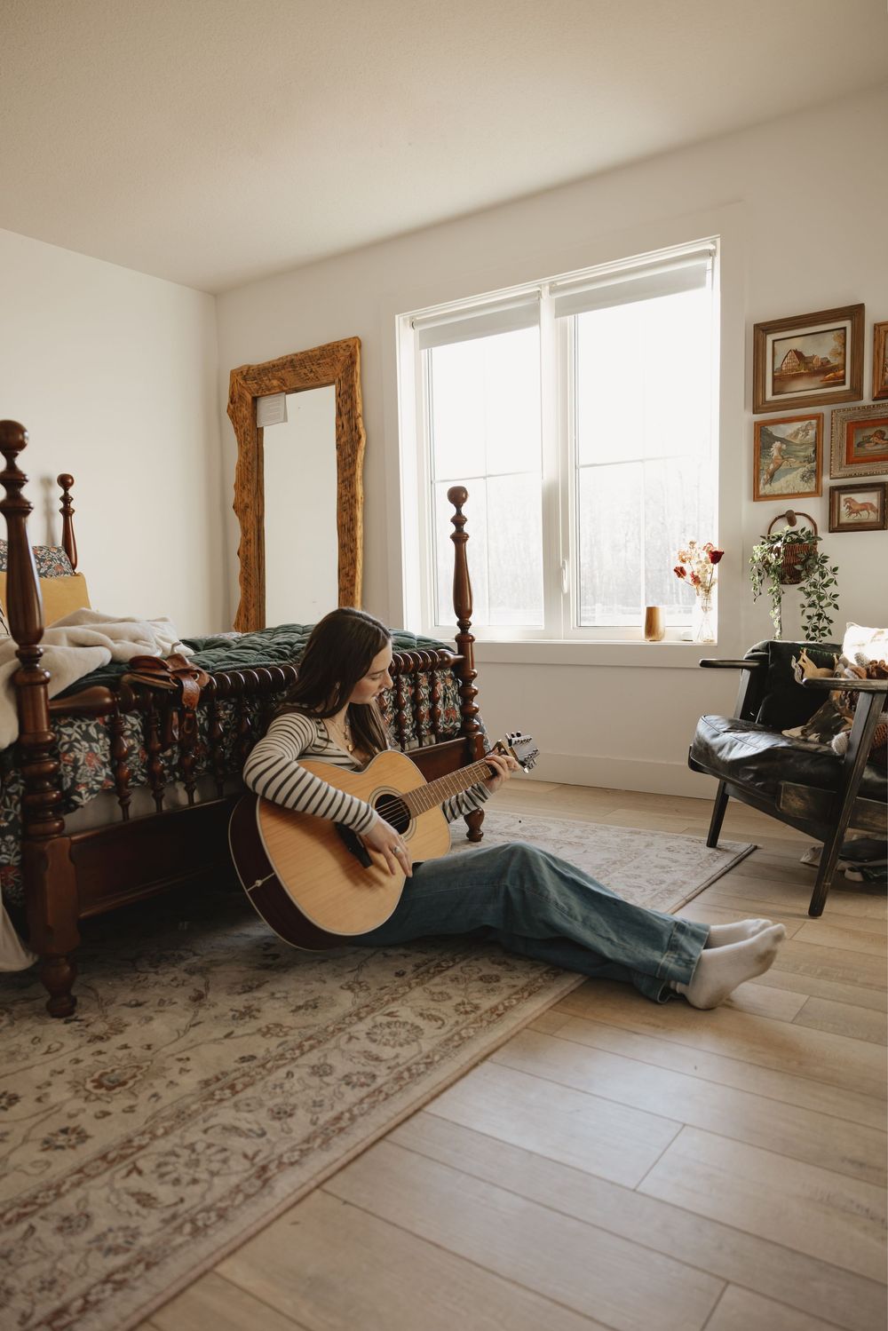 Young woman sitting on bedroom floor playing acoustic guitar, leaning against antique wooden bed with cozy decor.