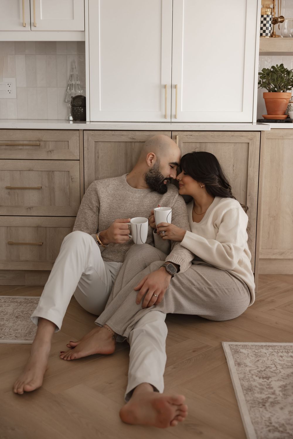 Couple sitting on kitchen floor in cozy neutral outfits, sharing an intimate moment while holding white mugs.