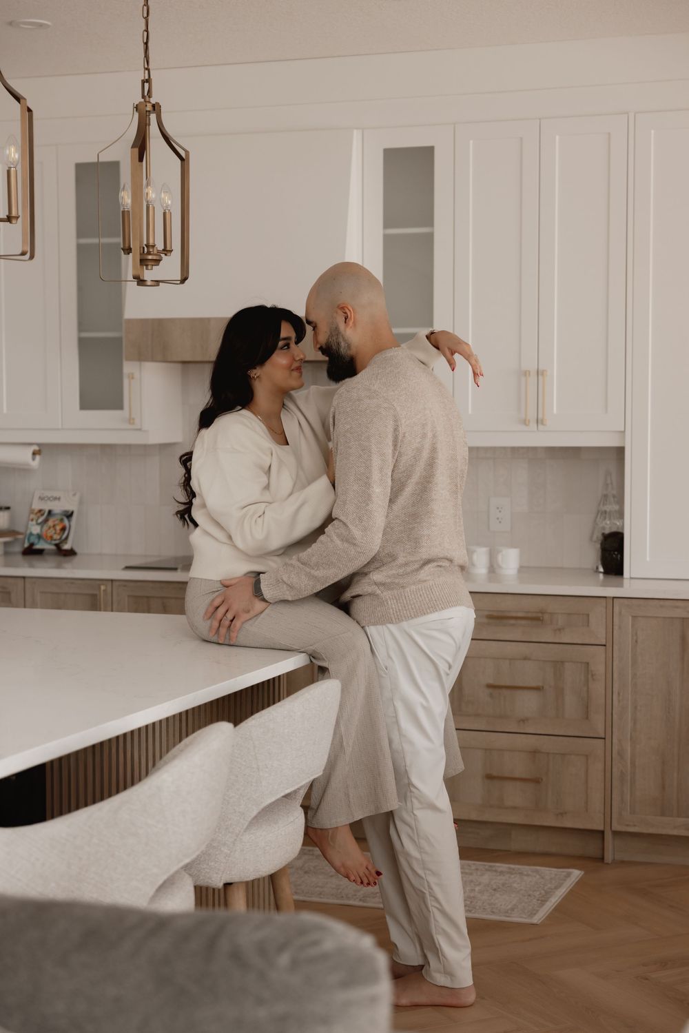 Couple sharing a romantic moment in a modern white and wood kitchen, woman sitting on counter embracing bald man.