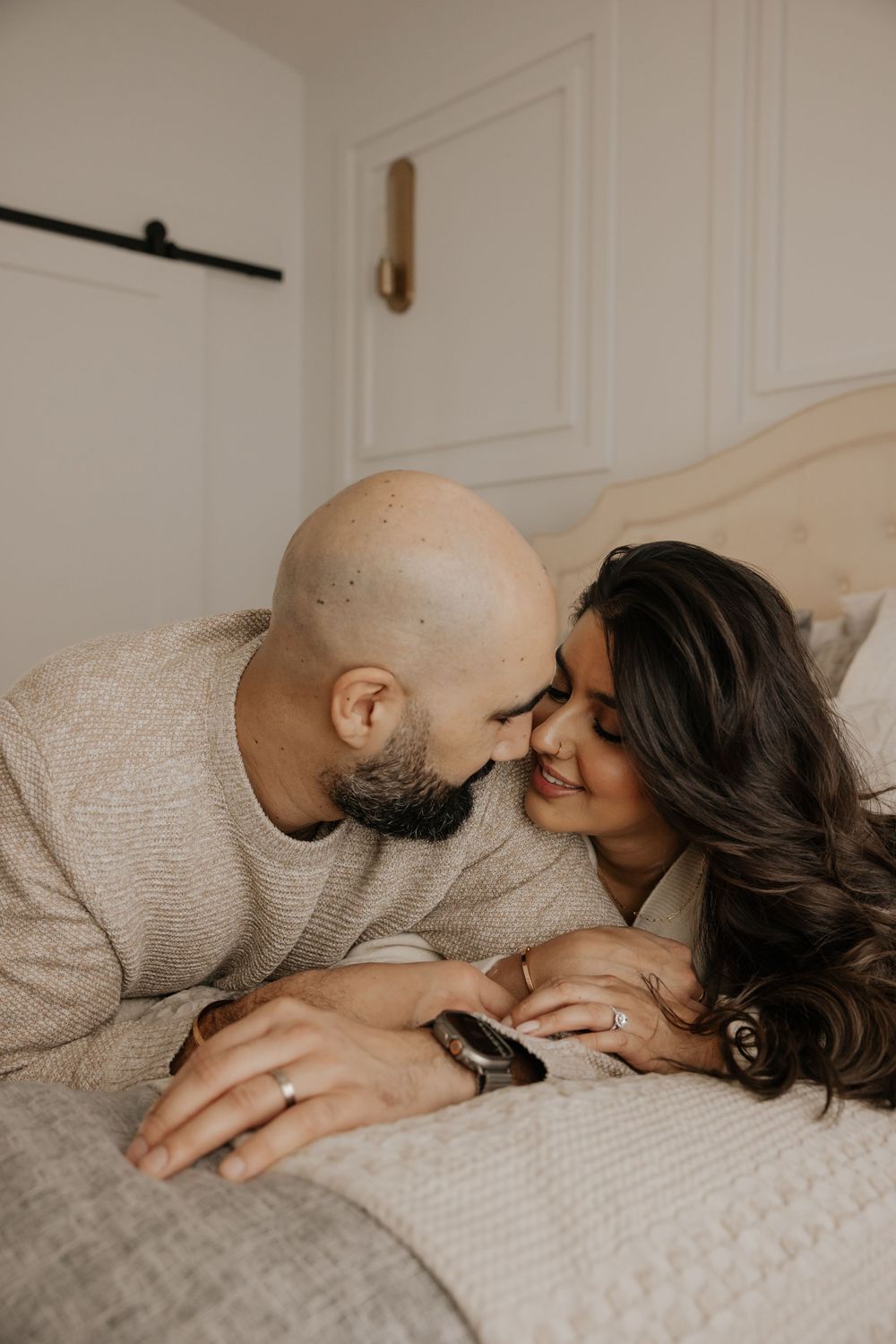 A couple shares a tender moment lying on a bed, the man leaning in to kiss the woman who smiles warmly, wearing an engagement ring.