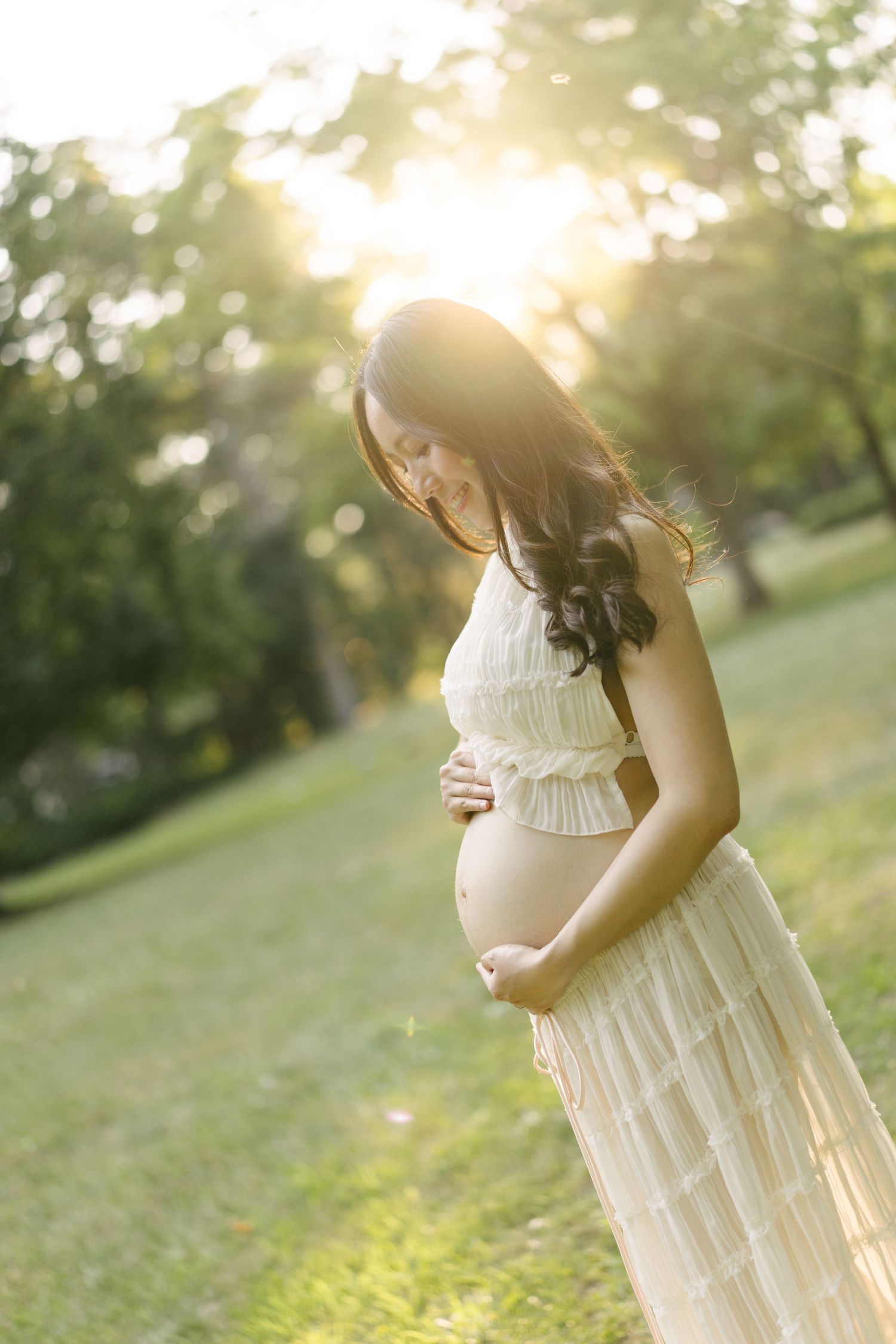 Expectant mother in a cream gown holding her belly in warm evening sun.