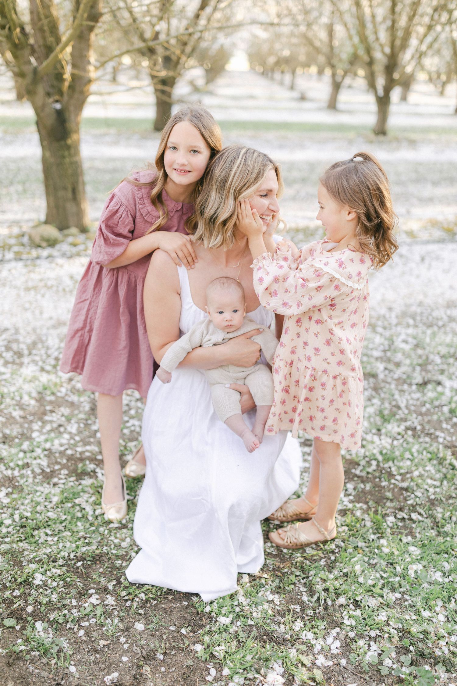 A mother in a white dress holds her baby while two young daughters in pink dresses embrace her in a blooming orchard.