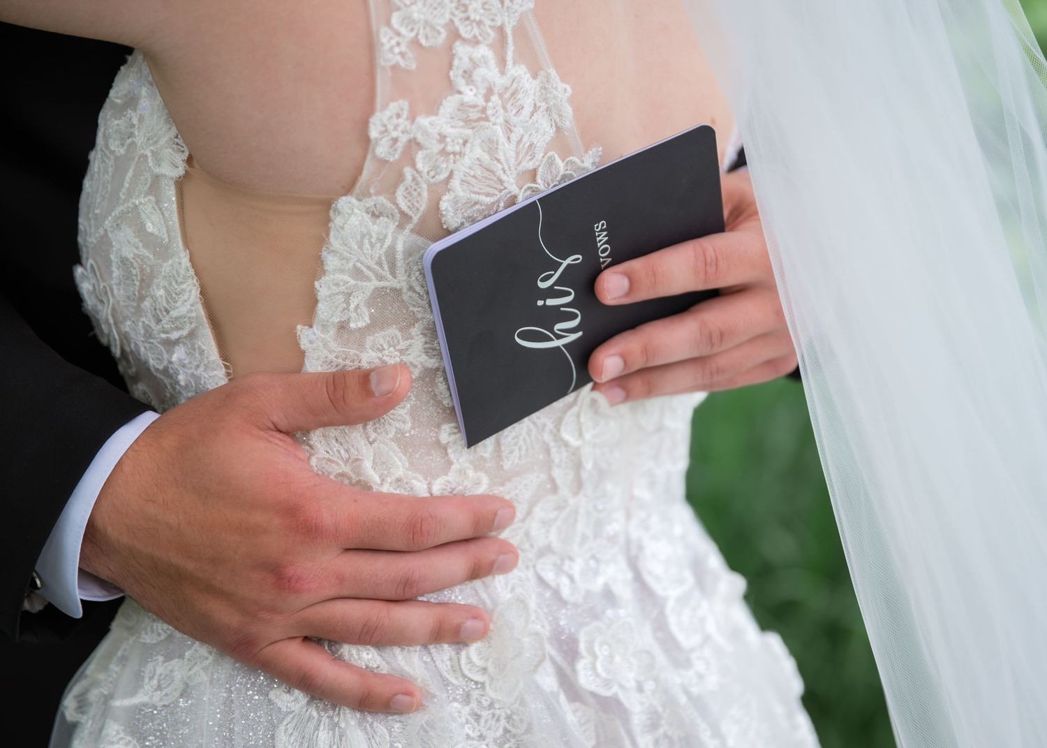 Bride in lace wedding dress holding a 'his' vow book while groom places hand on her back.