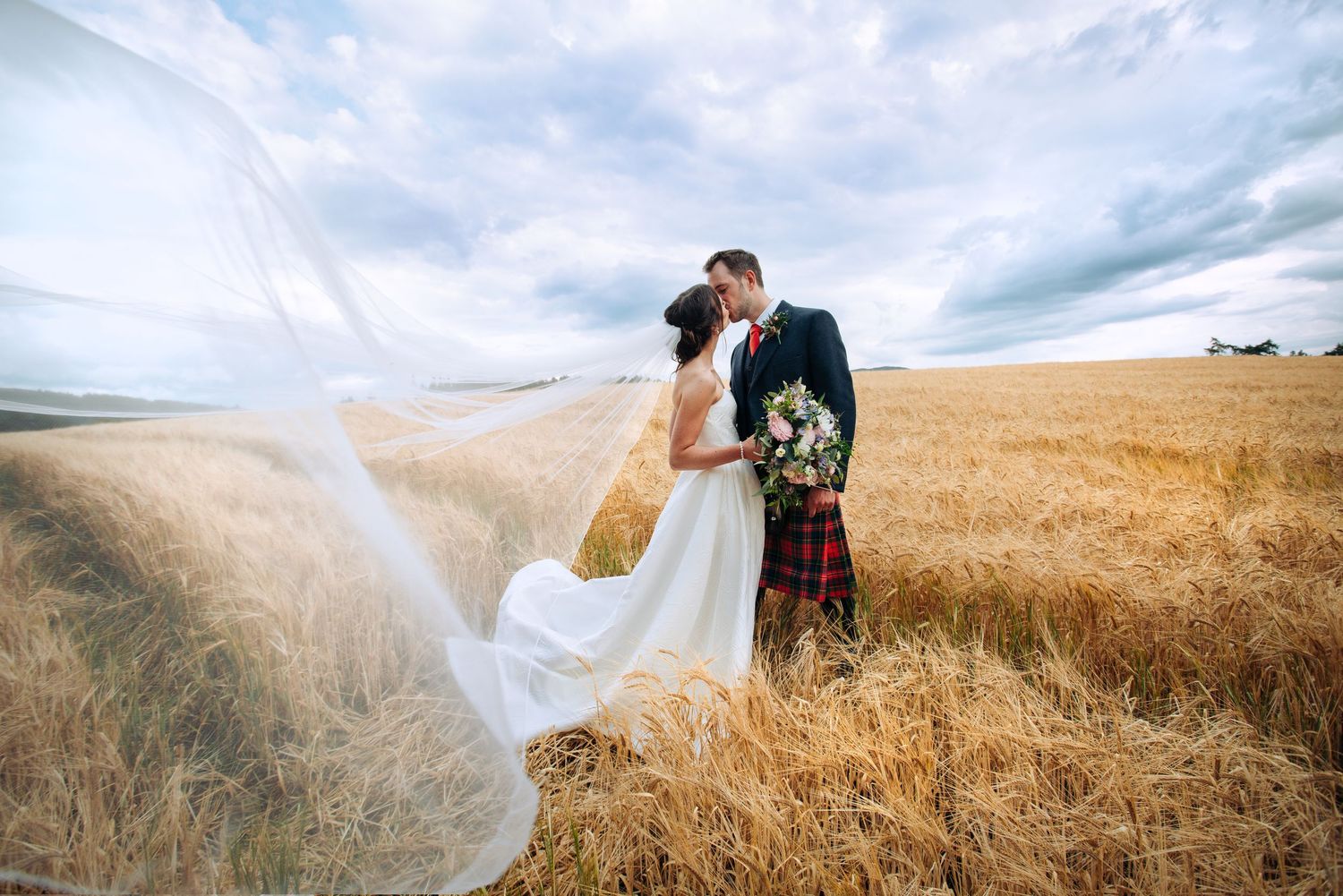 Bride and groom kissing in golden wheat field, veil flowing in wind, groom wearing Scottish tartan kilt.