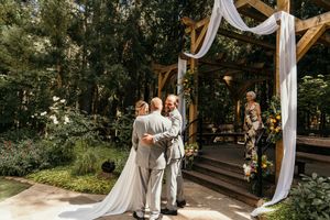 Outdoor wedding ceremony setup with white draped arch and wooden chairs in forest setting.