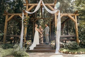 Elegant outdoor wedding ceremony setup with white fabric draped wooden arbors in a wooded forest setting.