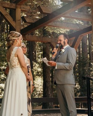 Wedding ceremony taking place under a wooden pergola in a sunlit forest setting.