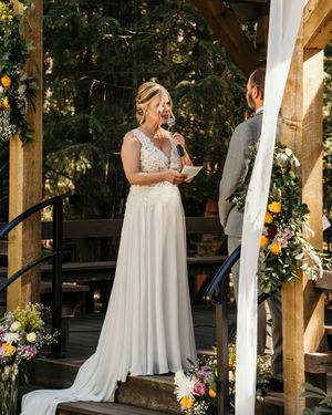 Wedding ceremony takes place under a rustic wooden pergola decorated with colorful flowers and white fabric.