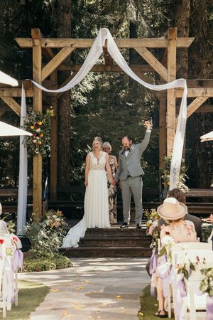 Outdoor wedding ceremony setup with wooden pergolas and white fabric draping in a wooded forest setting.