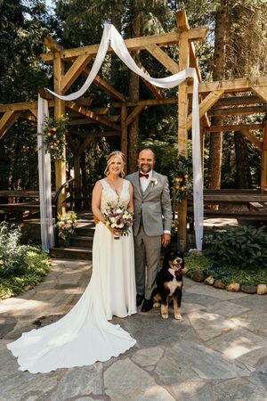 A wedding couple poses with their black dog under white draped arches in a rustic outdoor venue.