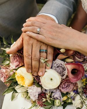 Close-up of hands with wedding rings resting on a lush floral bouquet with roses in pastel colors.