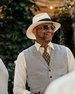A person in a light gray vest and white hat with sunglasses sits outdoors in natural lighting.