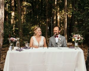 Long white reception table set up in a rustic forest setting with natural wood surroundings.