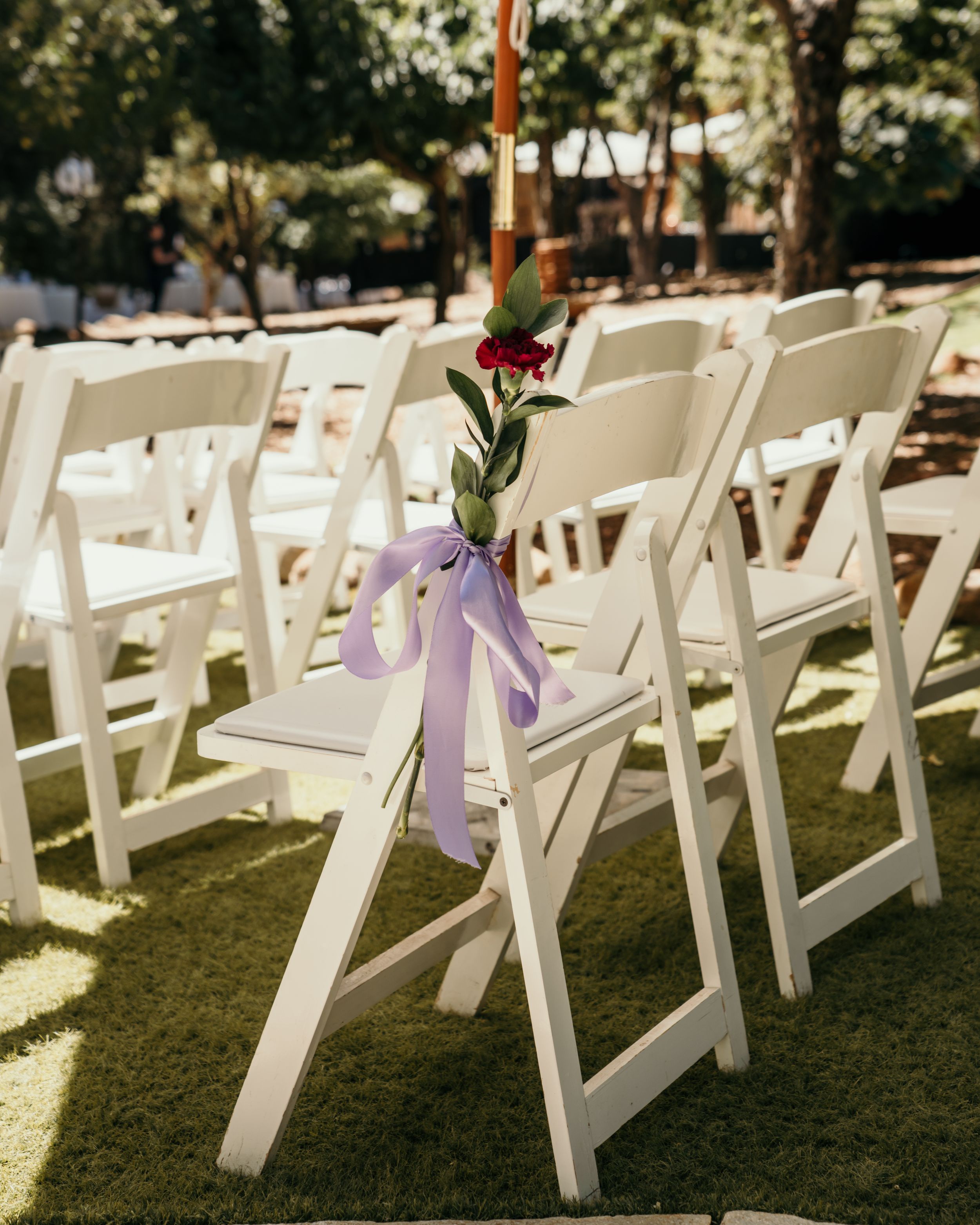 White folding chairs set up for an outdoor wedding ceremony with purple ribbons and floral aisle decorations.
