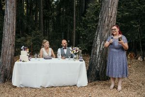 Wedding reception tables set up in a forest clearing with tall trees and string lights overhead.