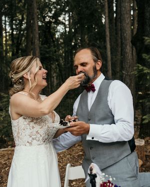Newlyweds share cake during their wedding reception in a forest setting.