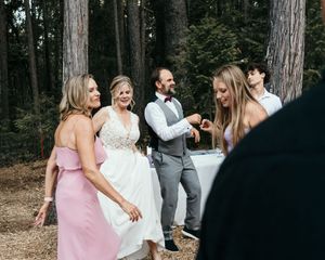 Wedding party celebrates and dances together in an outdoor wooded setting during golden hour.
