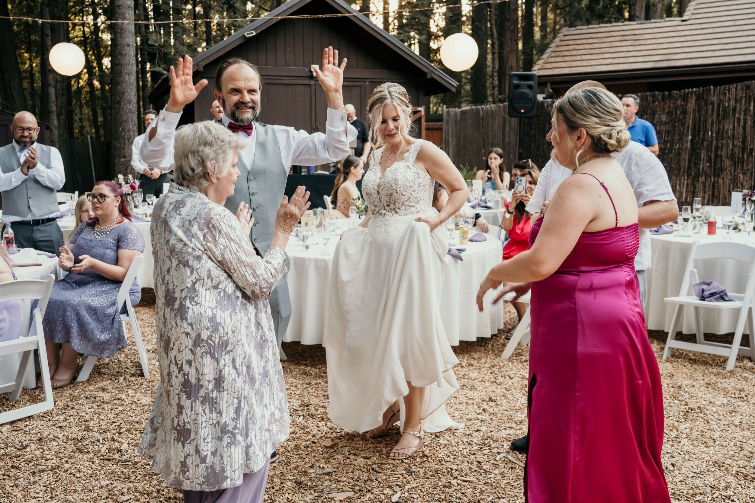 Wedding guests dancing and celebrating at an outdoor forest reception with string lights.