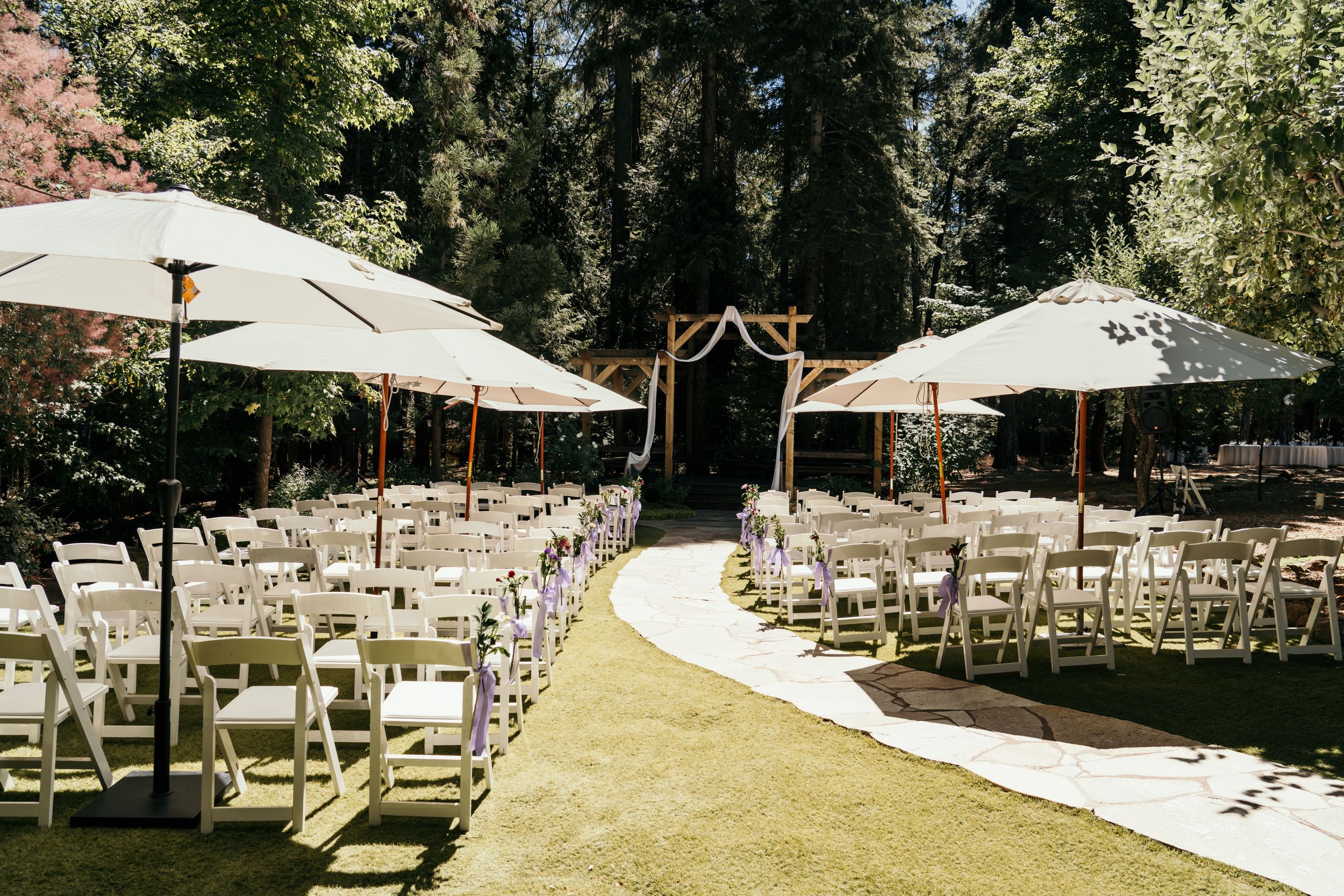 Outdoor wedding ceremony setup with white chairs and umbrellas arranged in rows under shady trees.