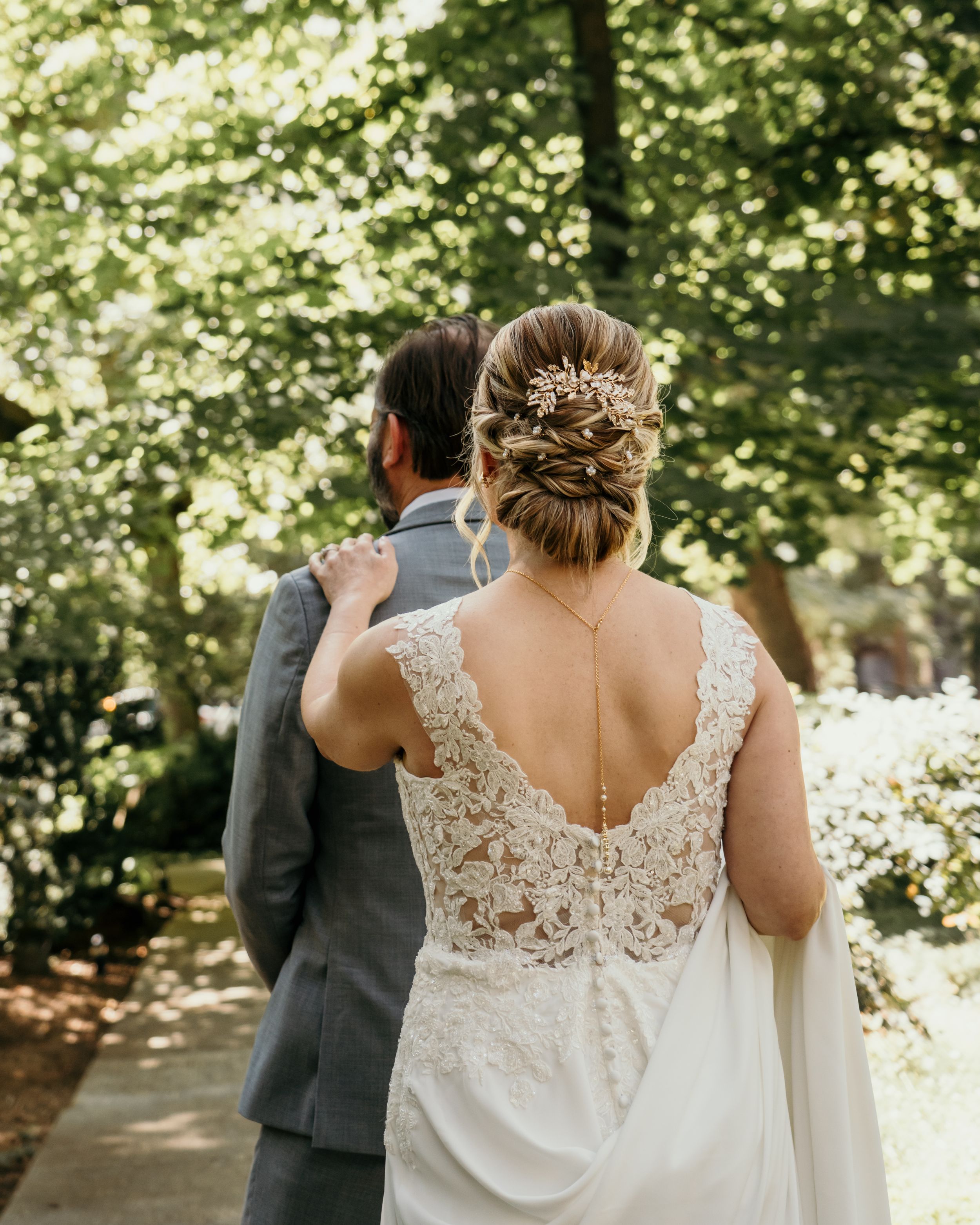 A couple in wedding attire walks through a sunlit wooded path with dappled light filtering through the trees.