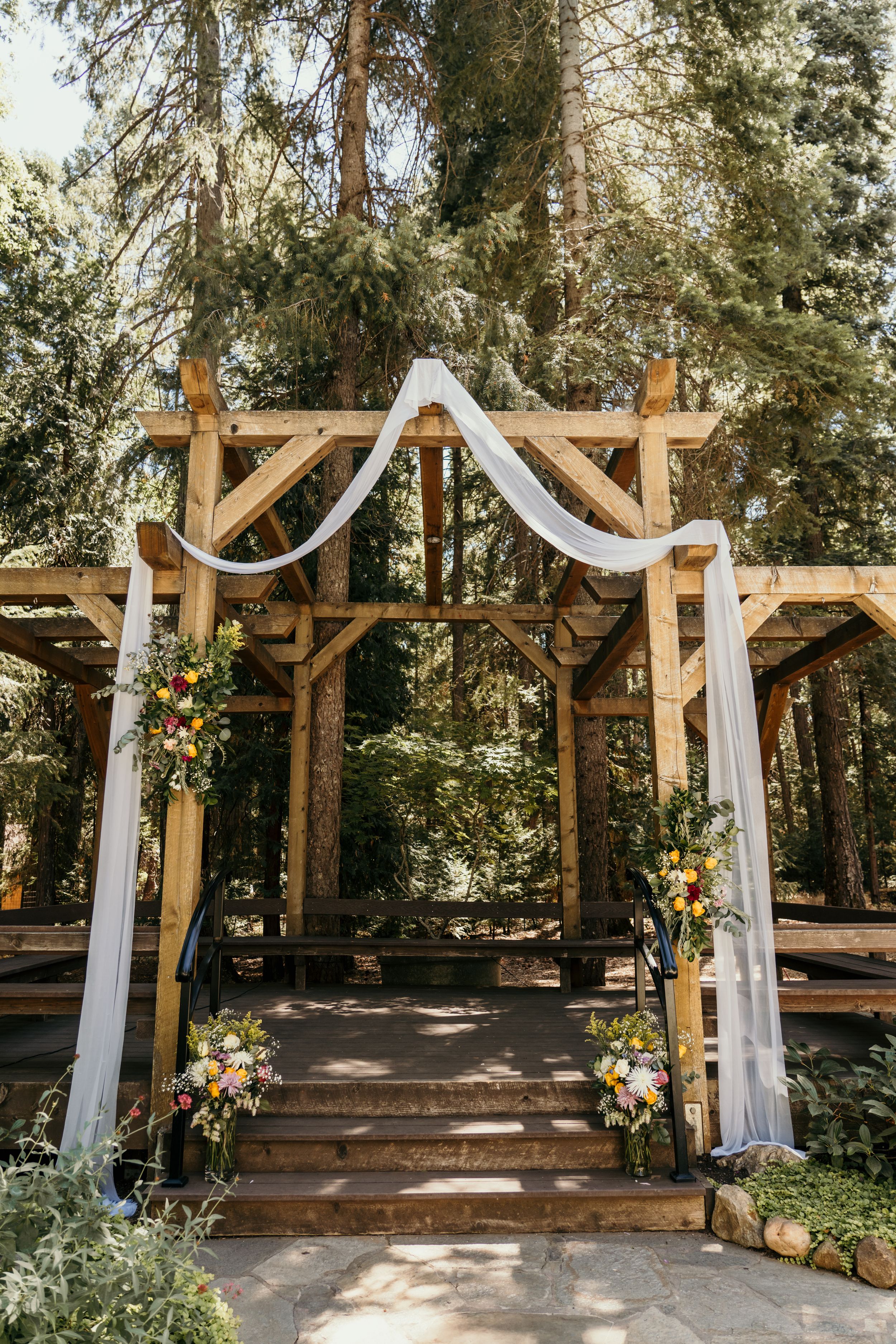 Rustic wooden wedding arch decorated with white fabric and floral arrangements in a serene forest setting.