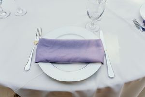 White place settings and lilac napkins arranged on elegant dining tables at a wedding reception.